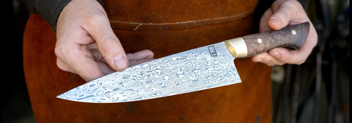 A close-up of a chef's knife with a patterned Damasteel steel blade and a figured Walnut handle with brass bolsters.
